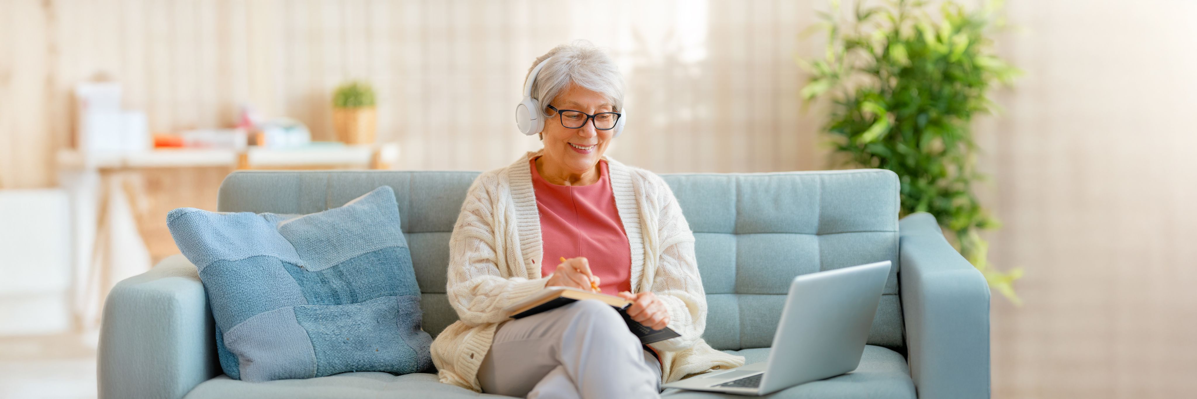 Photo of a woman sitting on a couch, looking at a laptop and taking notes in a notebook