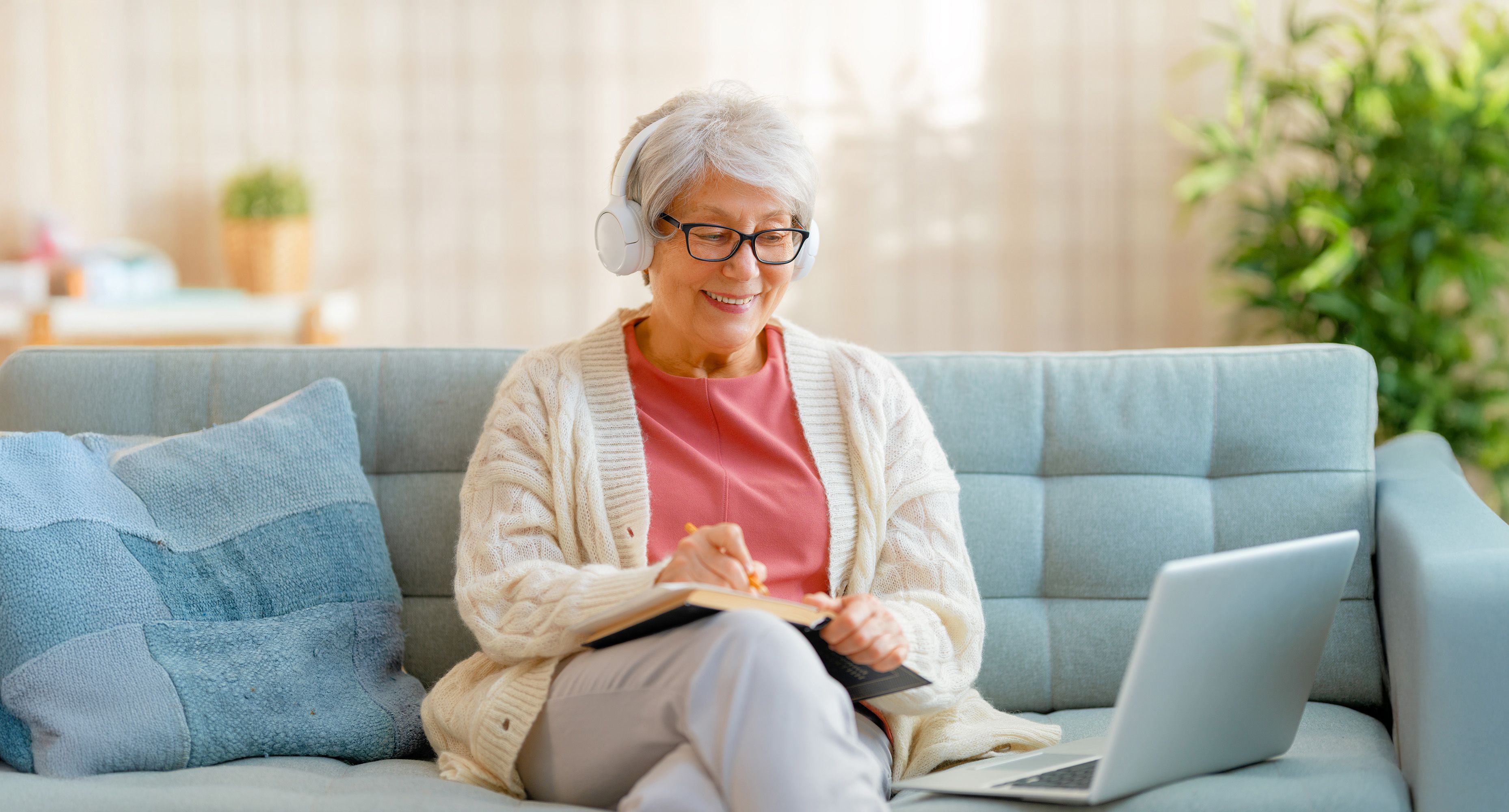 Photo of a woman sitting on a couch, looking at a laptop and taking notes in a notebook